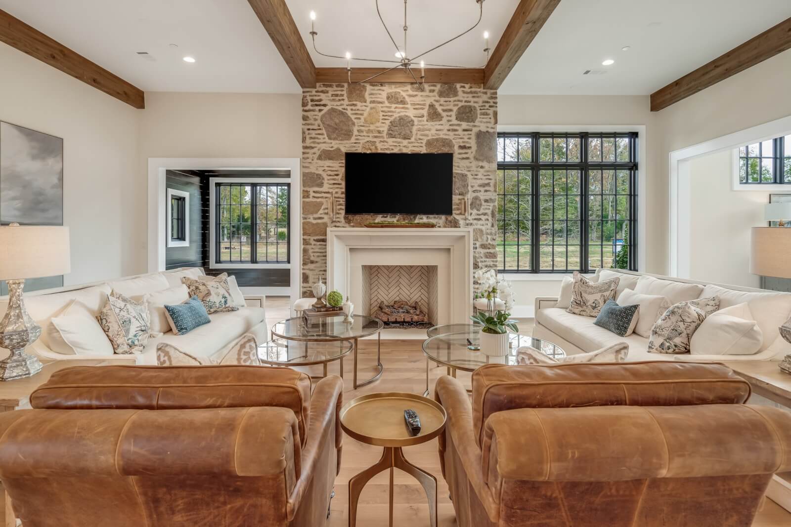 Incorporating natural stone, this living room features a fireplace beneath a mounted TV, complemented by two leather chairs, two sofas, and a round glass coffee table. Wooden beams grace the ceiling while large windows flood the space with natural light.