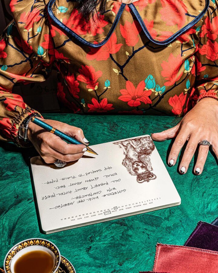 At a green table, a person in a floral blouse writes on holiday paper in their notepad, adorned with a pencil sketch of a lion. Nearby sit fabric swatches and a cup with saucer, creating an inviting scene.