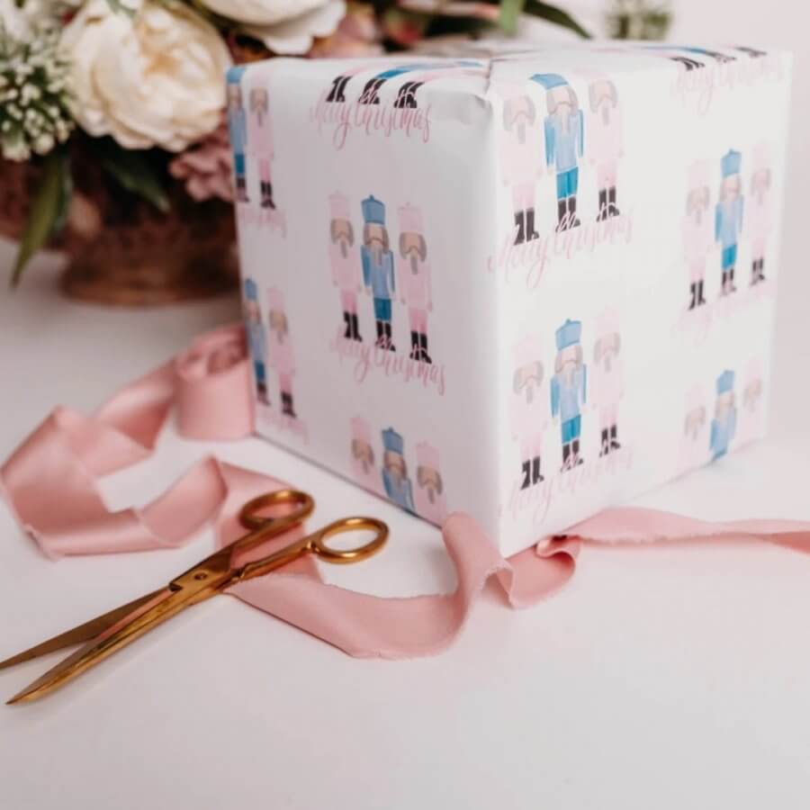 A gift box wrapped in festive Southern-made holiday paper, adorned with a pink ribbon, sits next to golden scissors on a white surface with flowers in the background.