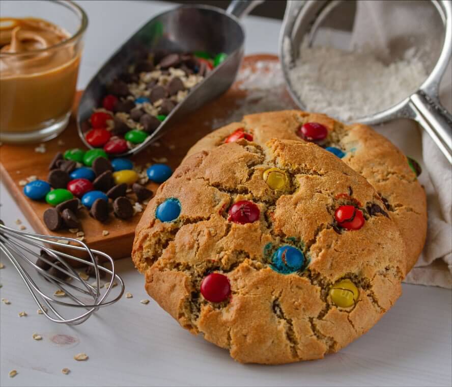 Two cookies with colorful candy pieces rest on a table, surrounded by baking essentials like peanut butter, chocolate chips, flour, and a whisk. Perfectly crafted by cookie companies that ship nationwide, these delightful treats bring sweetness right to your door.