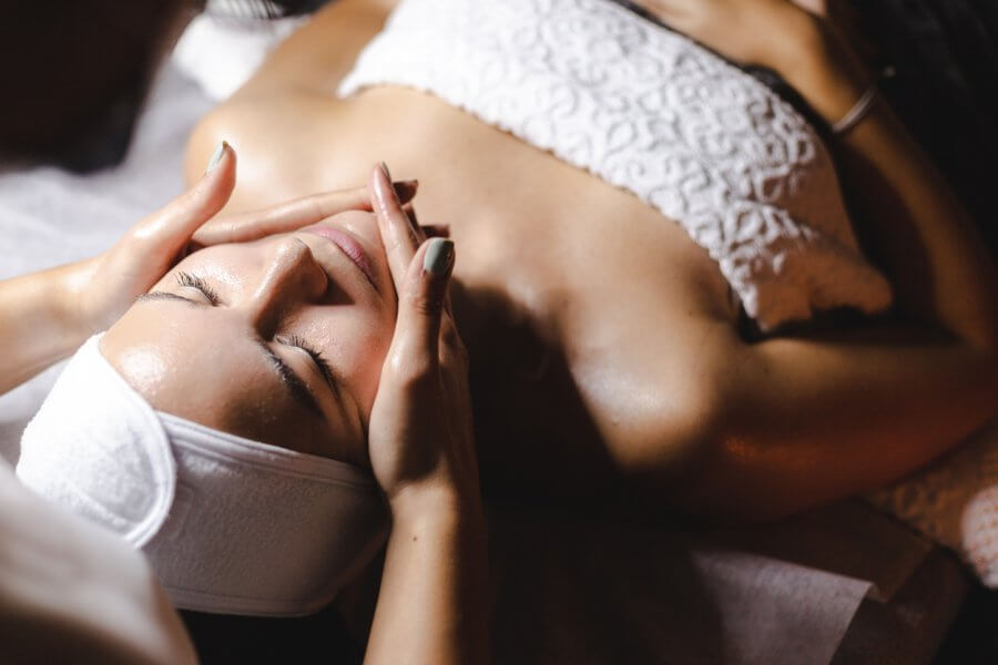 A person enjoys a soothing facial massage, lying down with closed eyes, wrapped in a white towel and headband. It's the perfect way to unwind and rejuvenate.