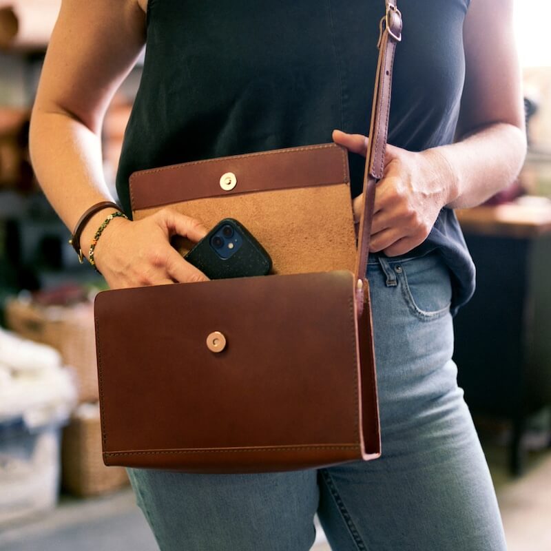 Standing indoors, a person carefully places a smartphone into an open brown leather crossbody bag