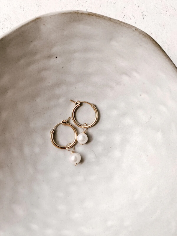 Gold hoop earrings, featuring pearl pendants, gracefully displayed on a textured white ceramic dish.