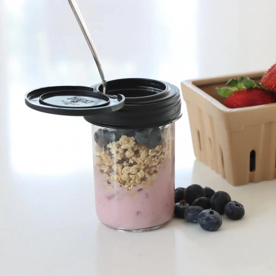 A jar with pink yogurt, granola, and blueberries, topped with a spoon. Nearby, a carton of strawberries and loose blueberries rest on a white surface.