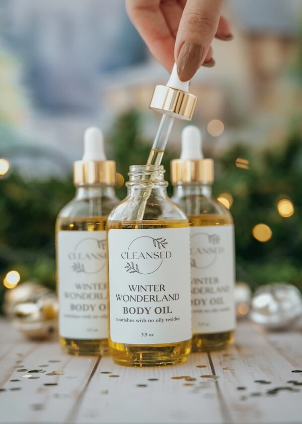 A dropper hovers over a bottle labeled "Winter Wonderland Body Oil," with two similar bottles in the background surrounded by festive decorations.