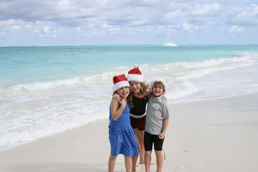 Three children wearing Santa hats stand together on a sandy beach with turquoise water and a boat in the background, embodying the joy of family trips during the holidays.