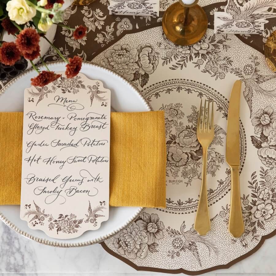 Elegant table setting with a handwritten menu wrapped around Southern paper, gold cutlery, mustard-colored napkin, and floral patterned plates. Perfect for holiday gatherings, the menu includes turkey, mashed potatoes, glazed carrots, and greens with bacon.