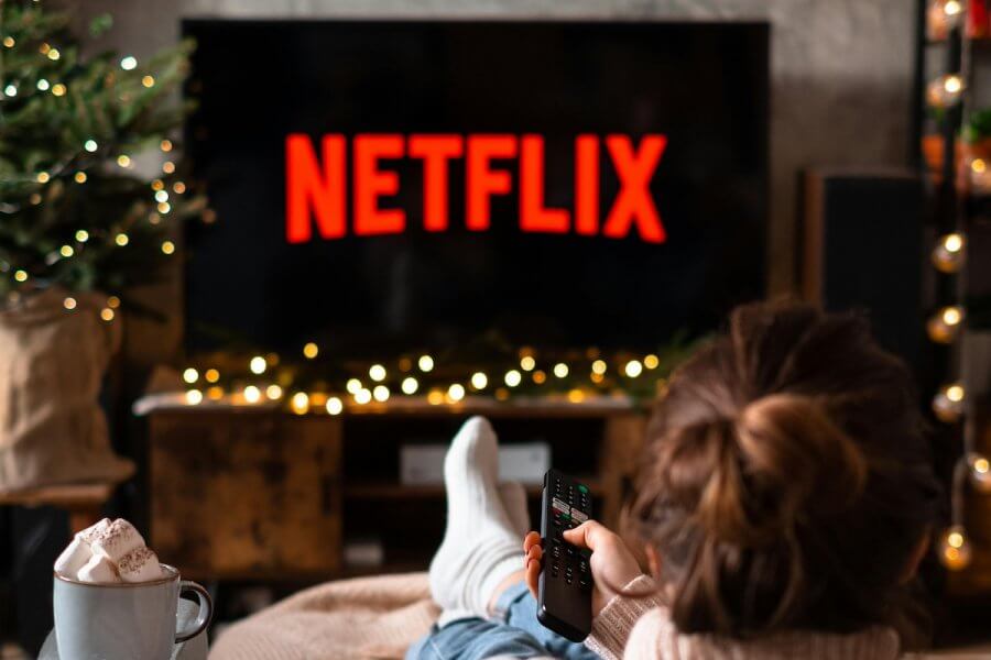 Person sitting on a couch, holding a remote, eyeing the TV displaying the Netflix logo. A mug of hot chocolate is on the table nearby as they settle in for a night with their binge-watch list.