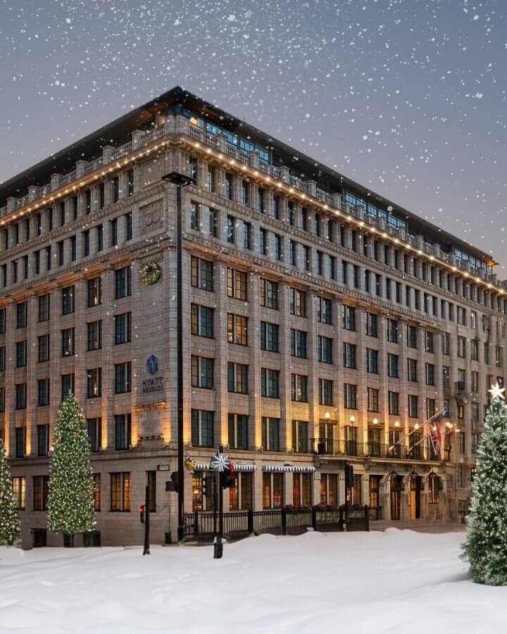 A large, multi-story hotel building is decorated with lights and surrounded by snow-covered trees. Snow gently falls from the sky.