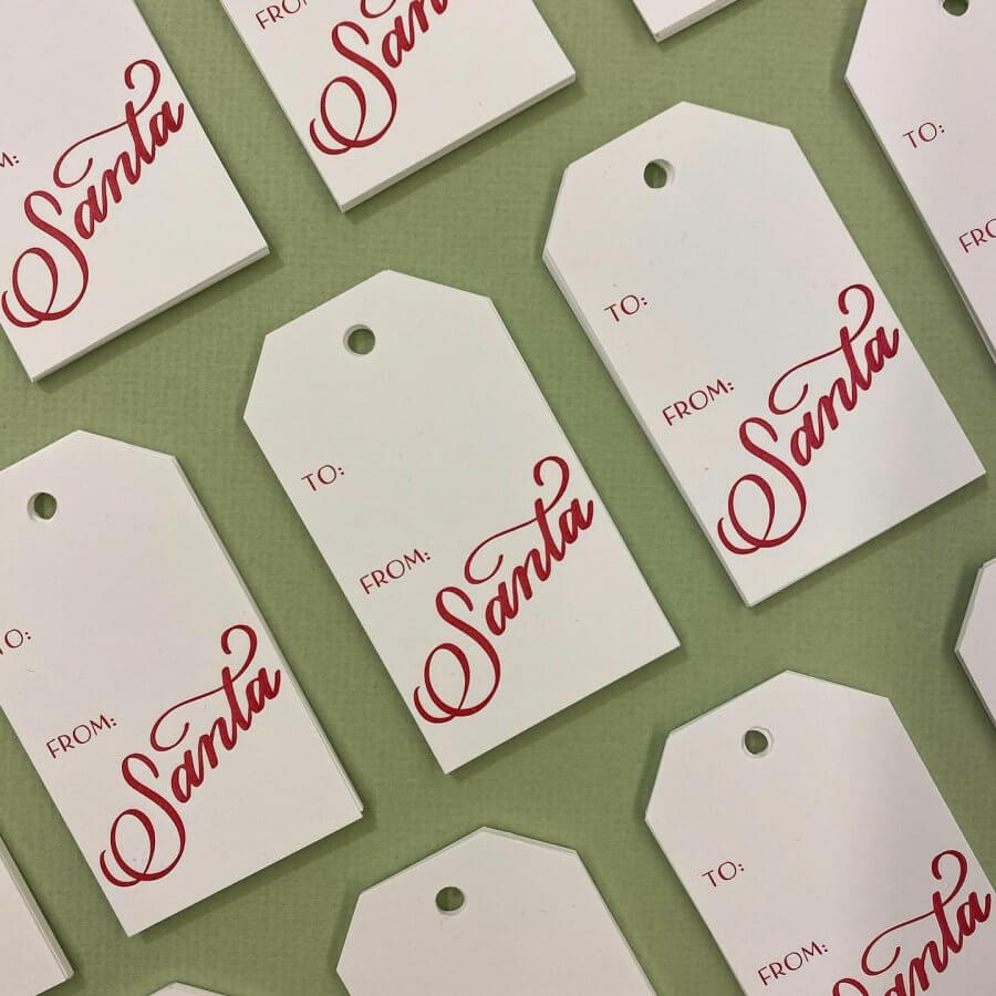 A collection of white gift tags with "To: From: Santa" written in red script, crafted by Southern makers, elegantly placed on a green surface. Perfect for adding a festive touch to your holiday wrap.