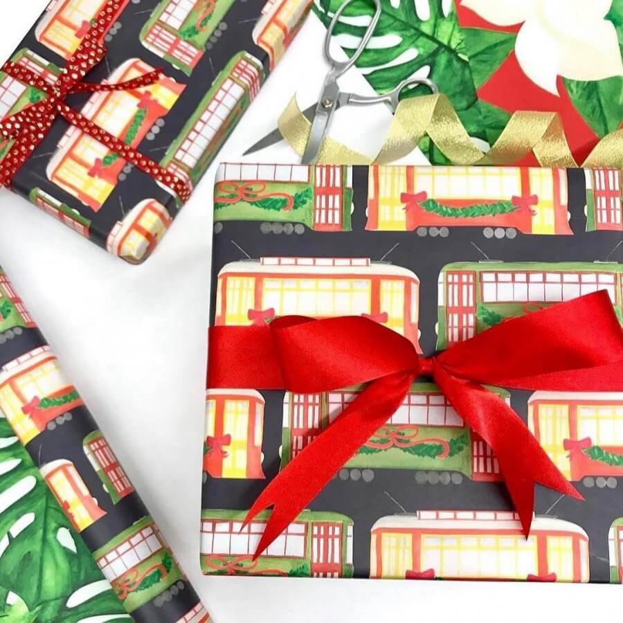 Gift boxes wrapped in trolley-themed paper, adorned with red and gold ribbons, surrounded by wrapping materials, scissors, and festive greenery—perfectly capturing the spirit of the holidays.