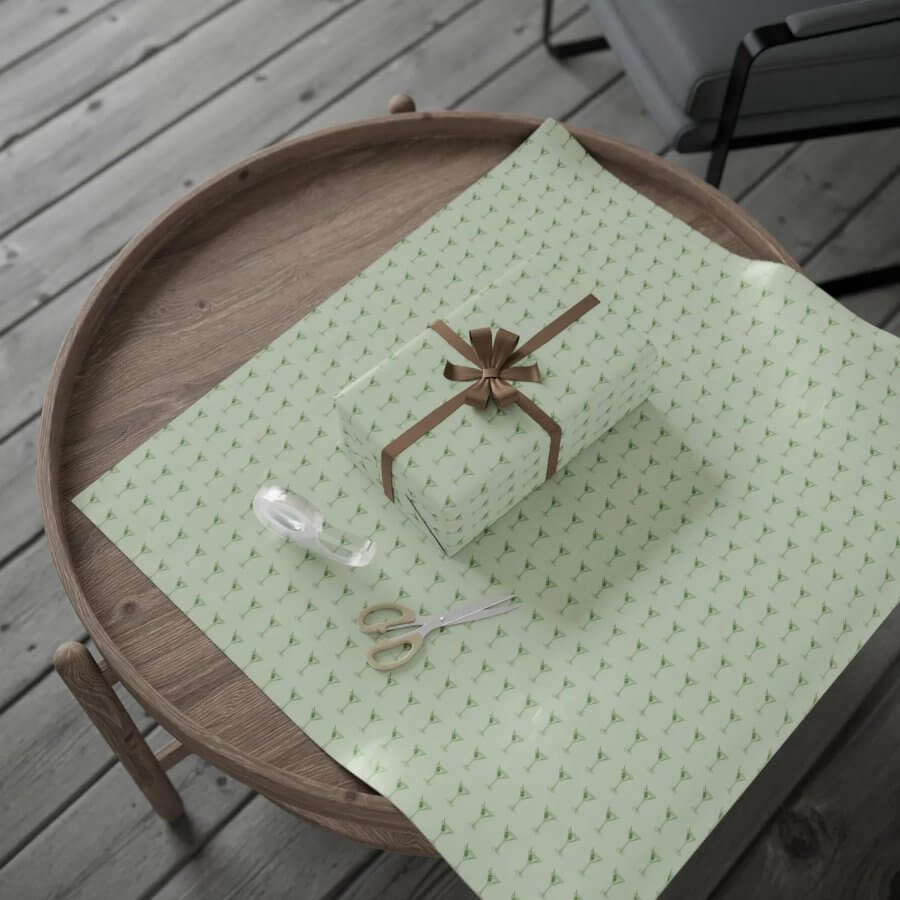 A wrapped gift with a brown ribbon sits on green patterned paper atop a round wooden table. Nearby are scissors and clear tape, ready for the holidays.