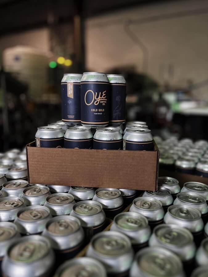 A cozy cardboard tray holding 12 cans of "Oye" Cold Gold beer sits atop a stack of similar finds in a Southern brewery setting.