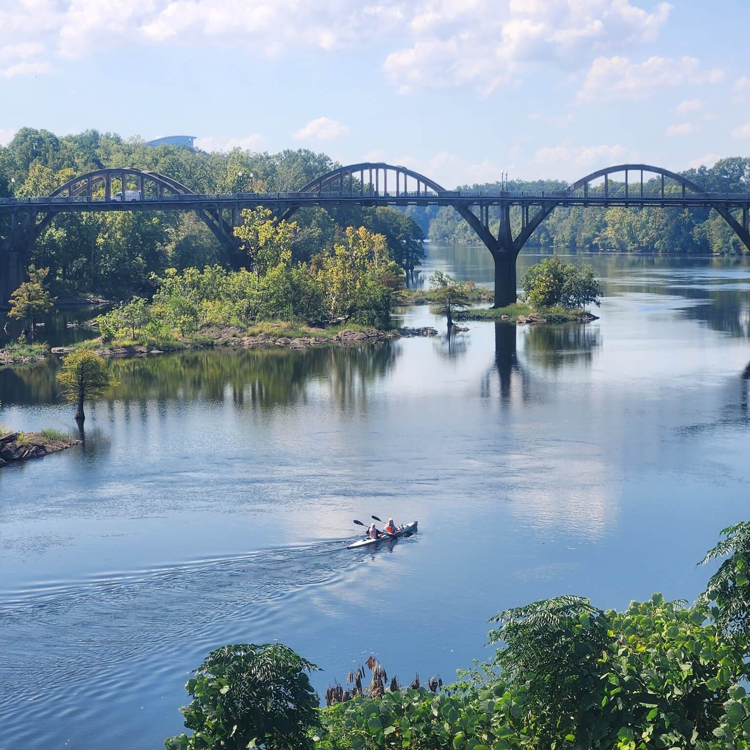 A small boat with rowers glides along a calm river under a bridge on a clear day, surrounded by lush greenery and small islands.