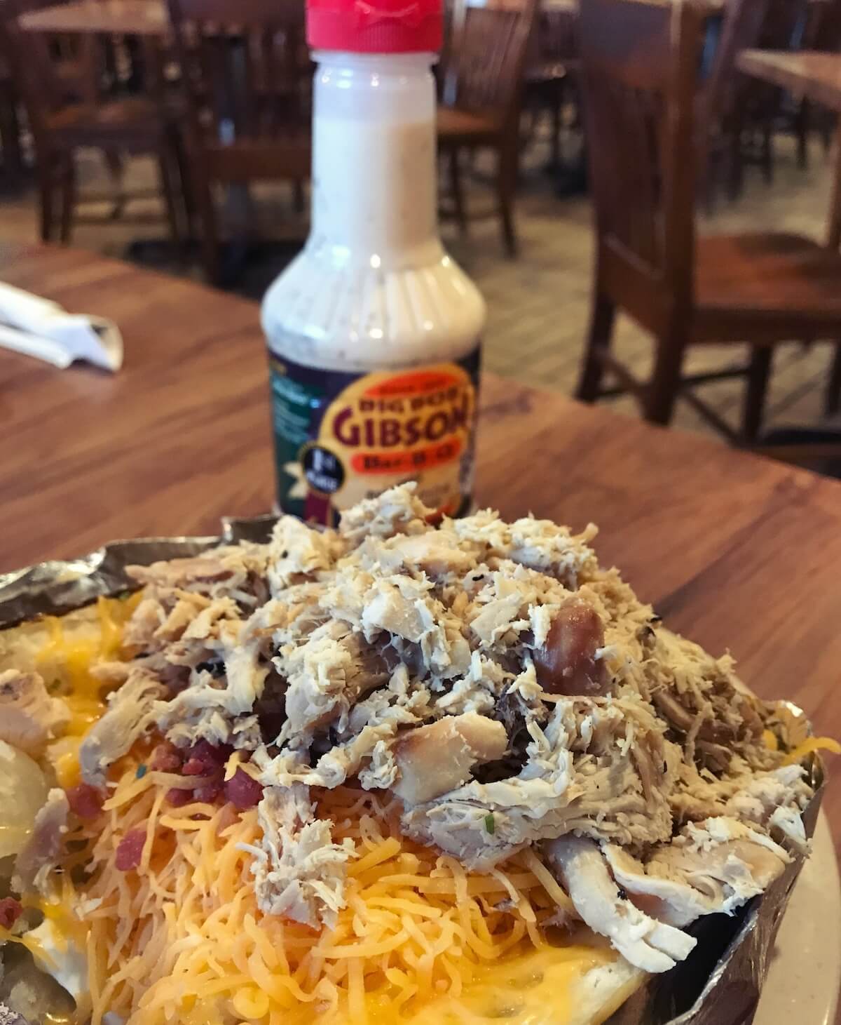 A loaded baked potato topped with shredded chicken, cheese, and bacon bits on a plate invites the flavors of one of the best towns to visit in Alabama. Nearby, a bottle of Big Bob Gibson's Alabama White Sauce sits on a wooden table, ready to enhance every bite.