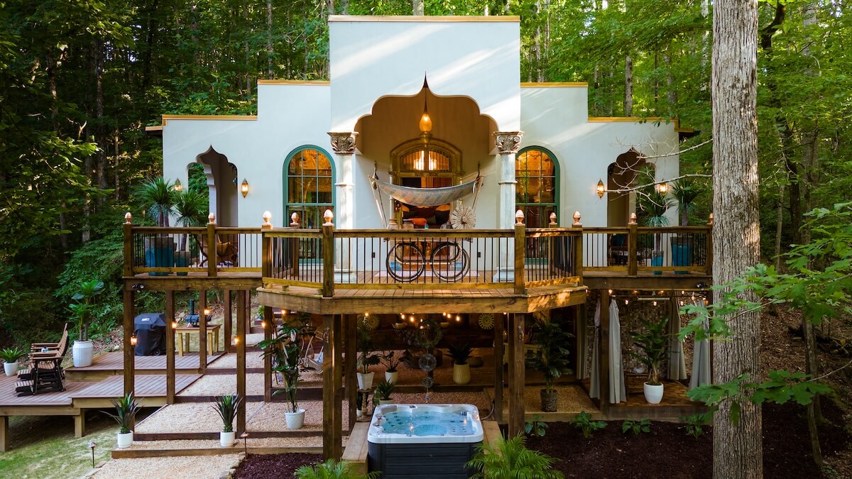 Nestled in a lush forest, this two-story house showcases unique architectural features like arched windows and a wooden deck. On the lower patio, surrounded by plants, a hot tub invites relaxation. It's the perfect retreat after exploring the best towns to visit in Alabama.