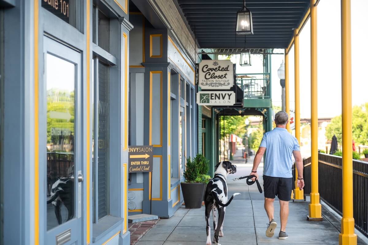 A man strolls along an Alabama sidewalk, a large black and white dog in tow, against the backdrop of charming shops. This picturesque scene captures the essence of travel, inviting you to explore new destinations and delightful local treasures.