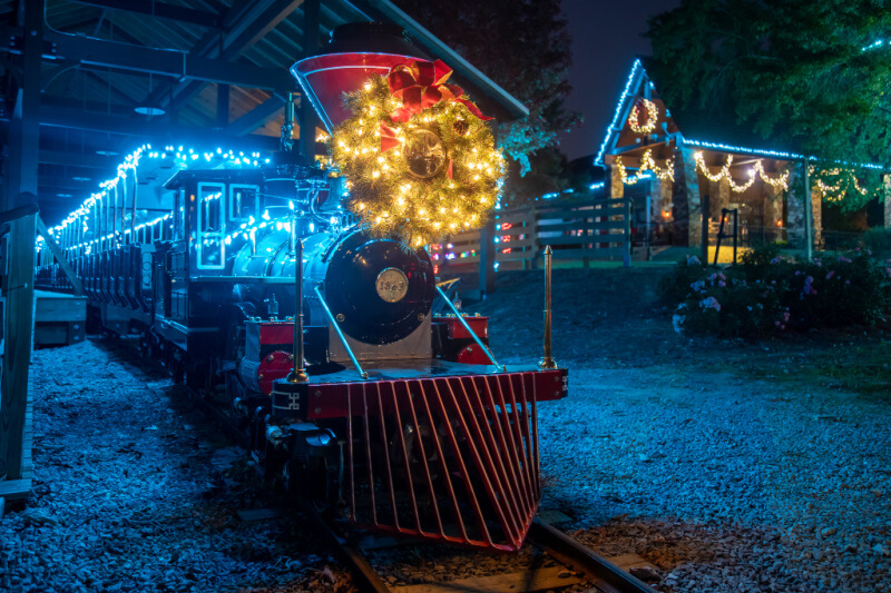 A festive, illuminated train with a glowing wreath on the front is parked in a decorated station at night, capturing the magic of Christmas in Alabama.