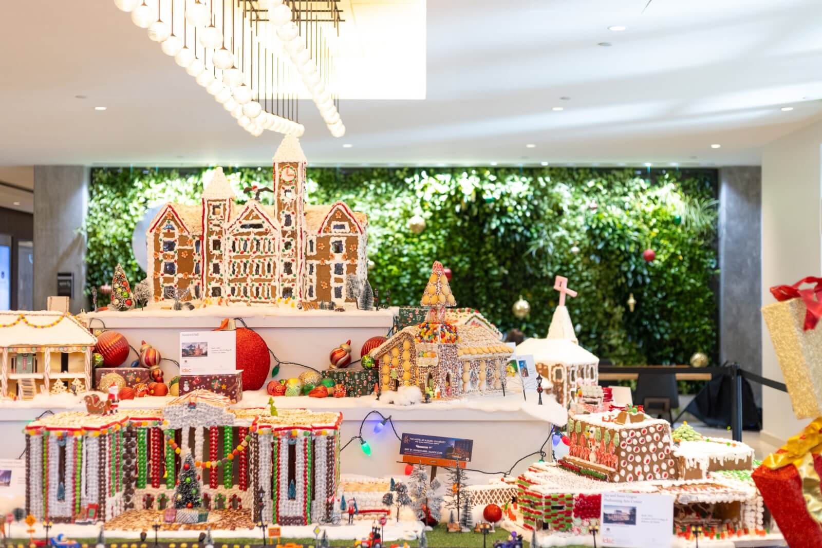 An elaborate gingerbread village display showcases various buildings adorned with festive decorations, all set against a backdrop of lush greenery. 