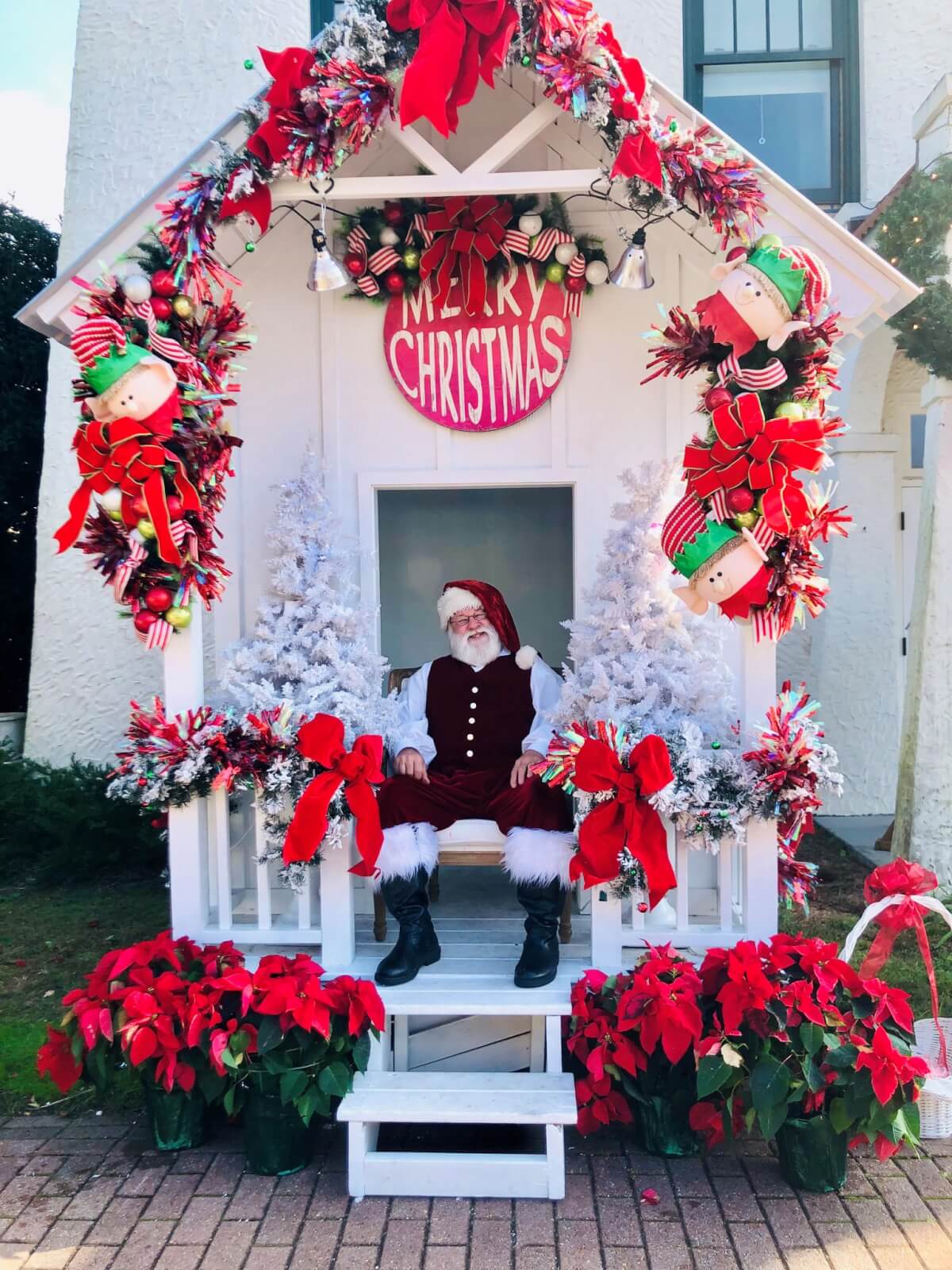 Santa Claus sits in a festive Christmas booth in Alabama, decorated with red bows, poinsettias, and white trees, making it one of the must-see holiday destinations.