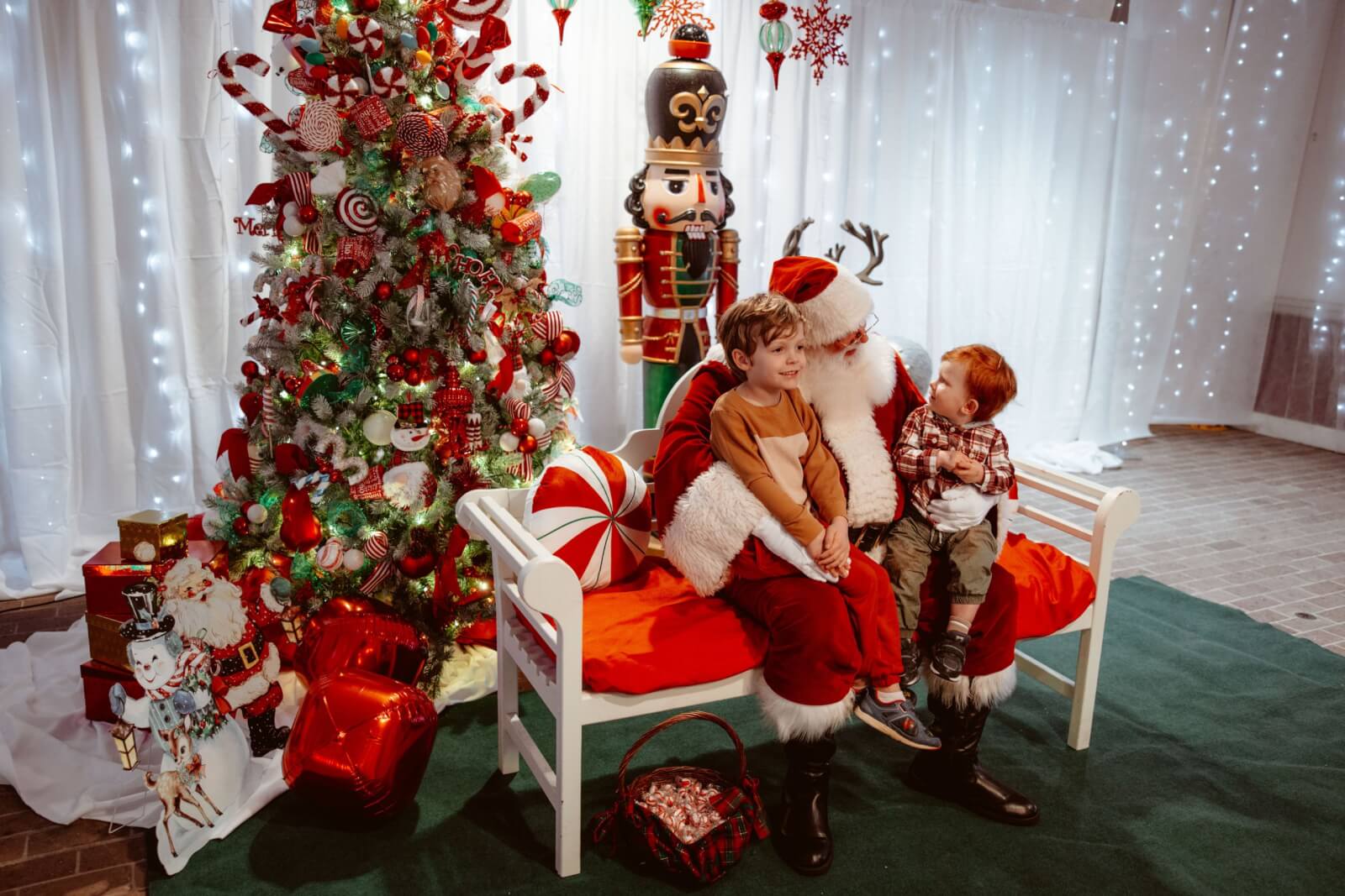 Two children sit with Santa Claus on a bench, creating a charming holiday scene that could make Alabama proud. With a Christmas tree and a large nutcracker nearby, it captures the magic of cherished holiday destinations.