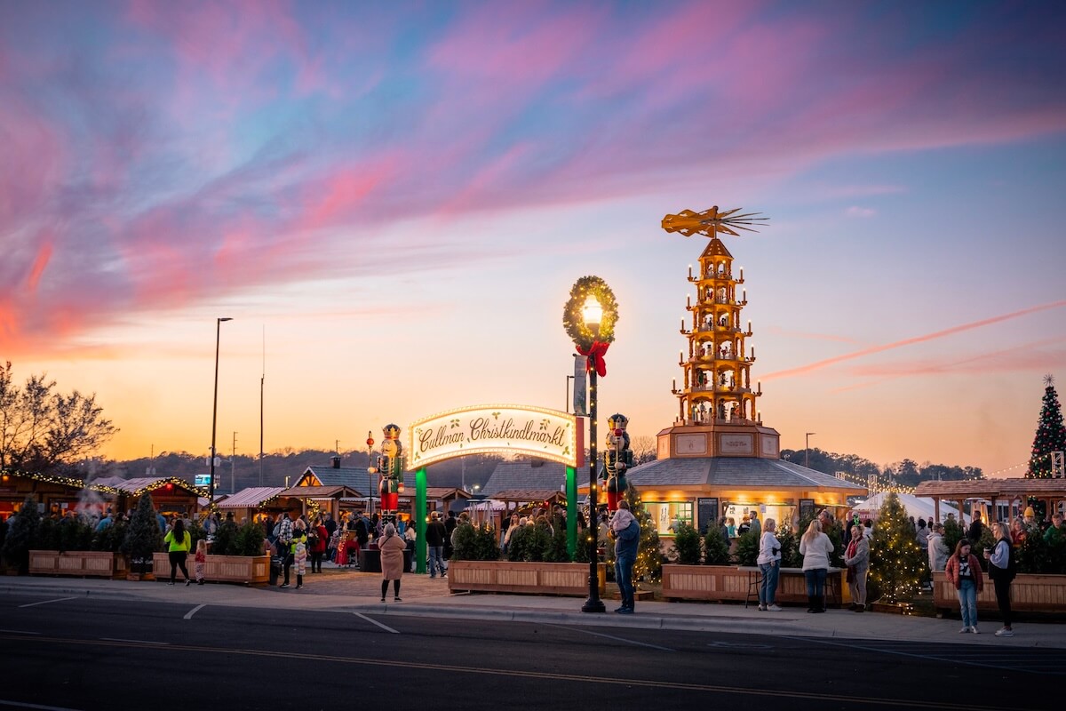 At sunset, people enjoy a Christmas market in one of the charming towns in Alabama, featuring festive lights, a large wooden pyramid, and beautifully decorated trees.