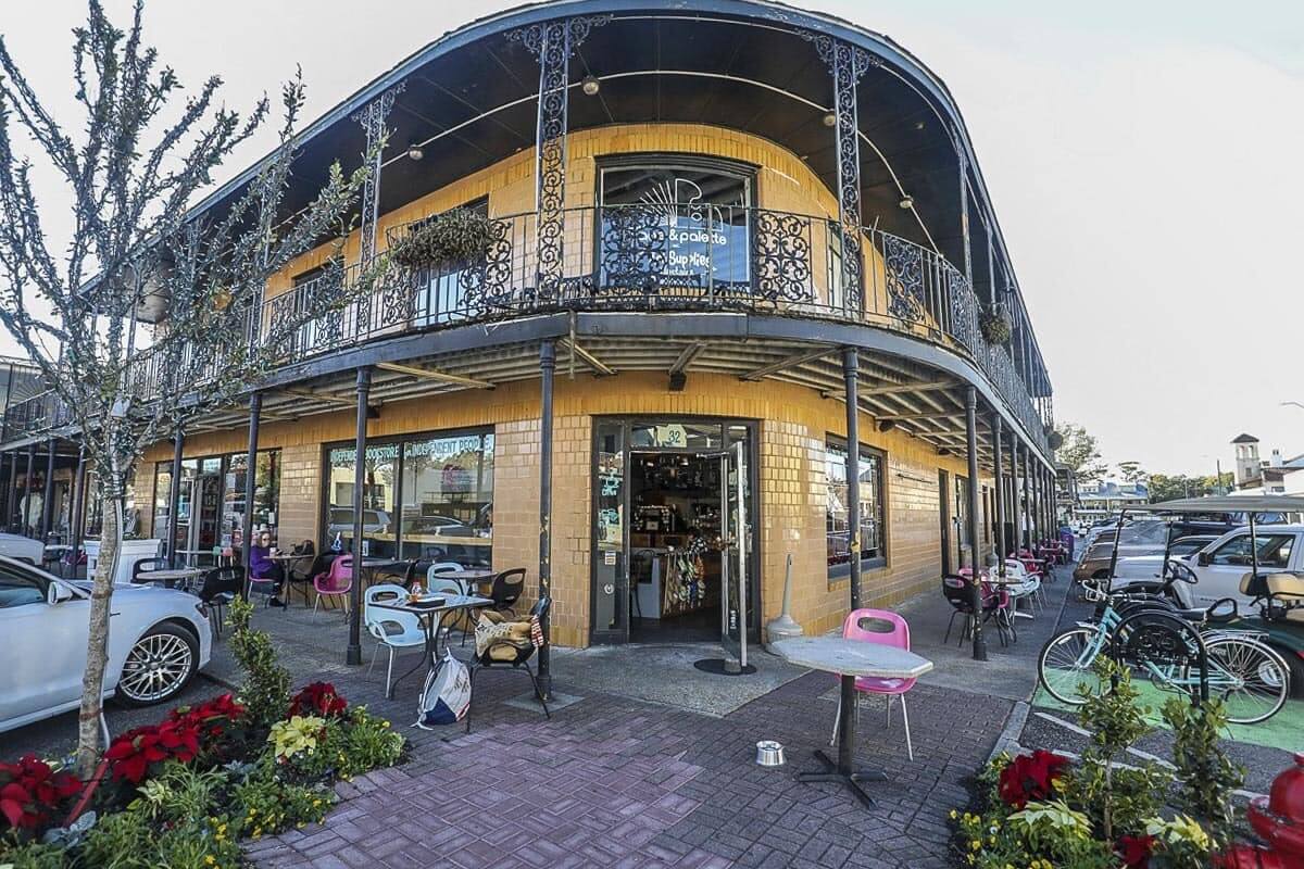 This corner café and bookstore features outdoor seating under a decorative iron balcony. Various chairs and tables dot the sidewalk, surrounded by potted plants and parked bicycles, creating a charming Southern ambiance.