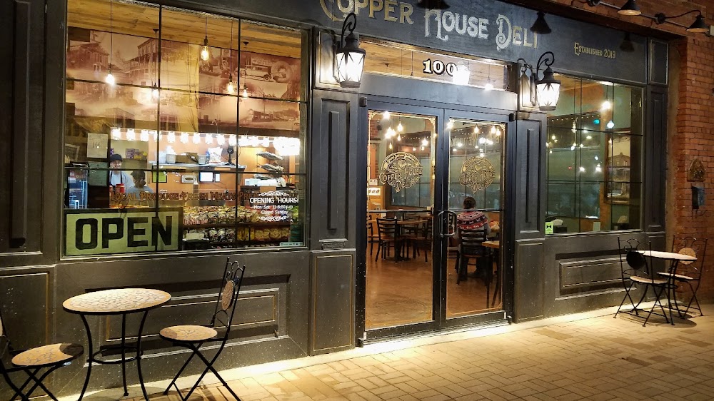 At night, the Copper House Deli stands out like a cozy gem among towns in Alabama, with two tables and chairs outside and an inviting open sign on the window. The warmly lit interior is beautifully visible through the glass doors, welcoming all who pass by.