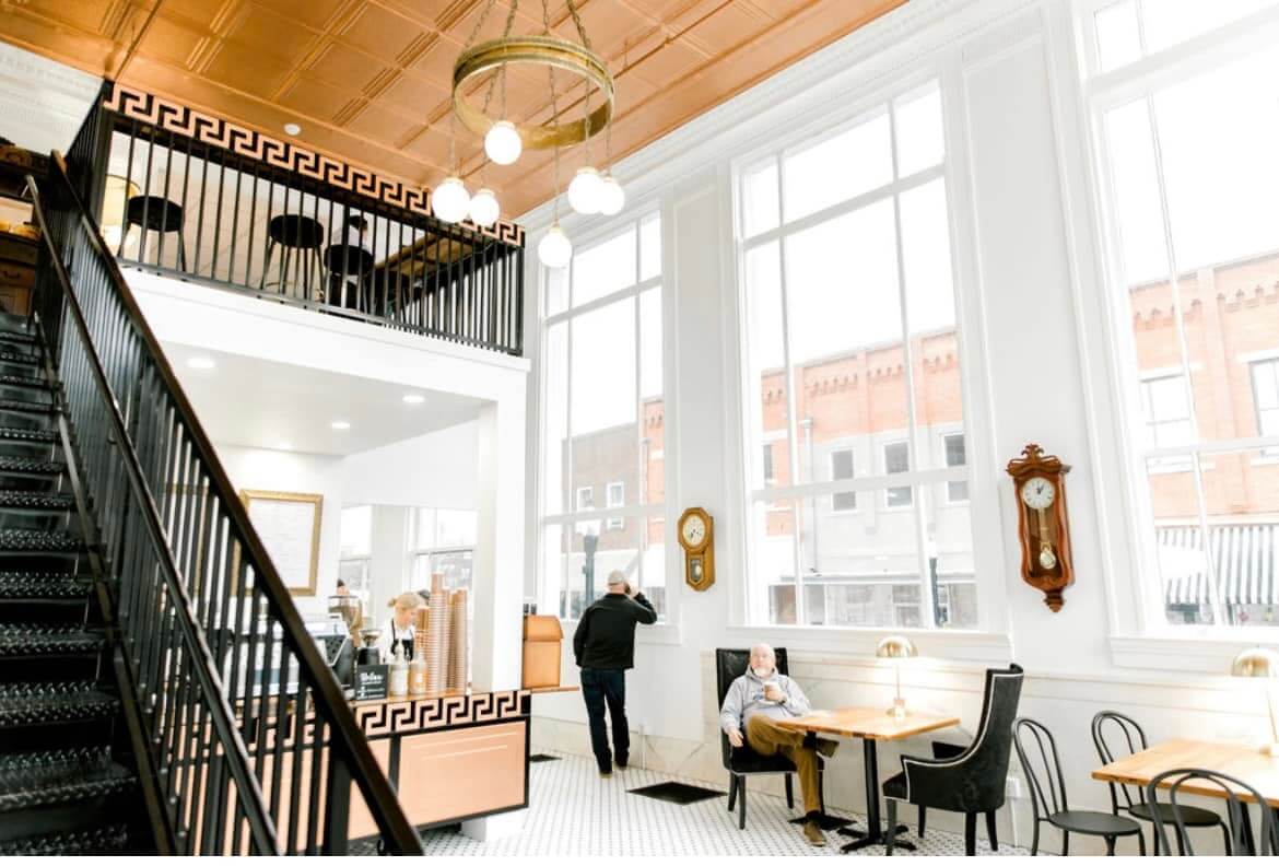 A bright, modern cafe with high ceilings and large windows, reminiscent of charming towns in Alabama, features people seated at tables and a staircase leading to an inviting upper level.