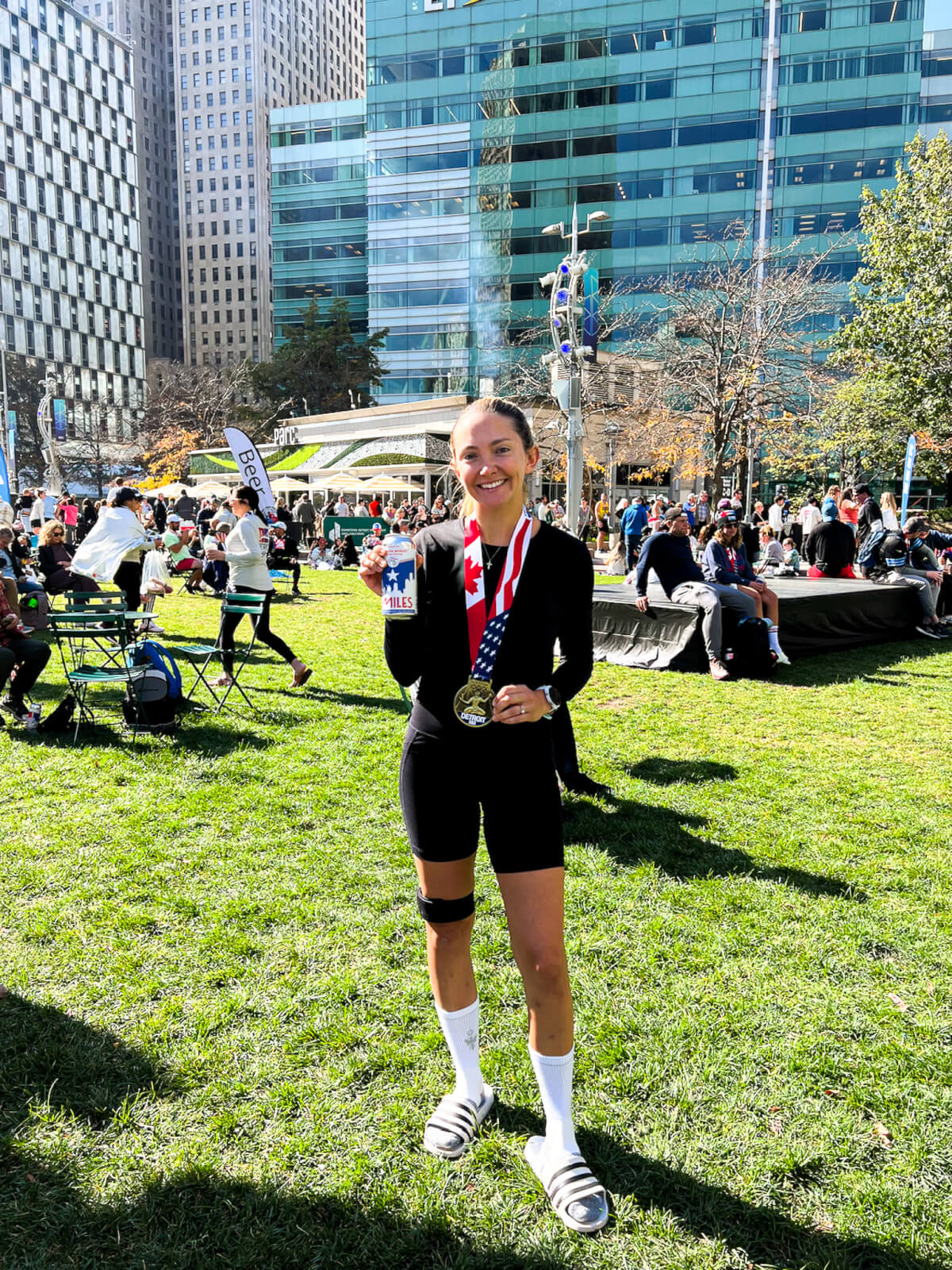 A person in athletic gear stands on the lush grass, proudly holding a medal and a water bottle, embodying the triumph of completing one of the best spring marathons. In the background, people mingle among picturesque buildings on a sunny day.