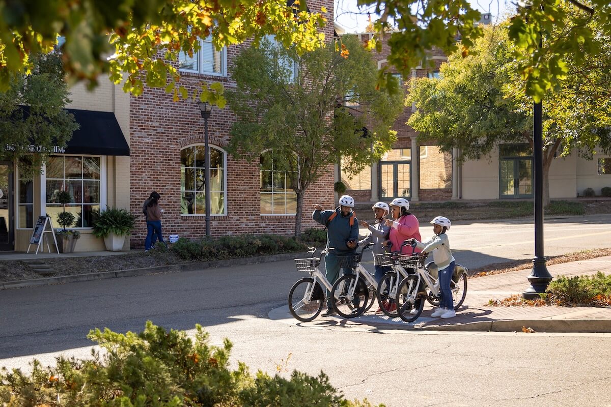 A group of four people on bicycles stops at a street corner in Ridgeland, with trees and brick buildings in the background.