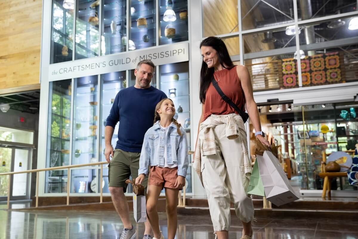 A family of three strolls joyfully through the Craftsmen's Guild of Mississippi in Ridgeland, their shopping bags full and smiles wide—just one of the many reasons to visit MS.