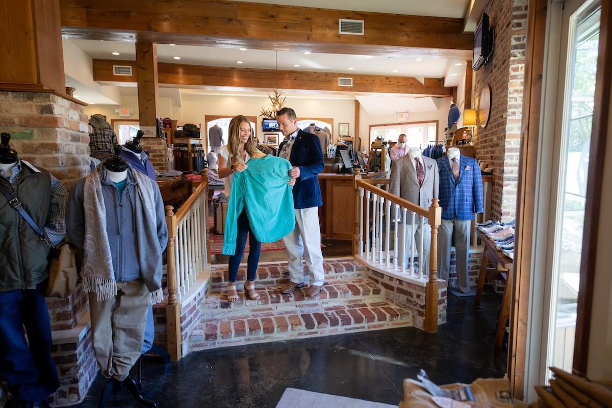 A woman and man look at a turquoise jacket in a Ridgeland clothing store, where various outfits are displayed against wooden beams and brick walls. One of the many reasons to visit this charming MS spot is its unique blend of style and Southern charm.