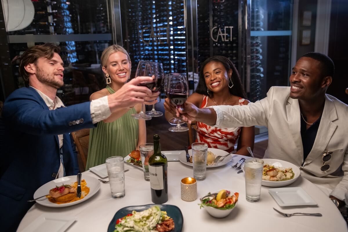 Four people seated at a restaurant table in Ridgeland, MS, clink wine glasses over plates of delicious food. A bottle of wine and water glasses are visible as they celebrate their visit to this delightful dining spot.