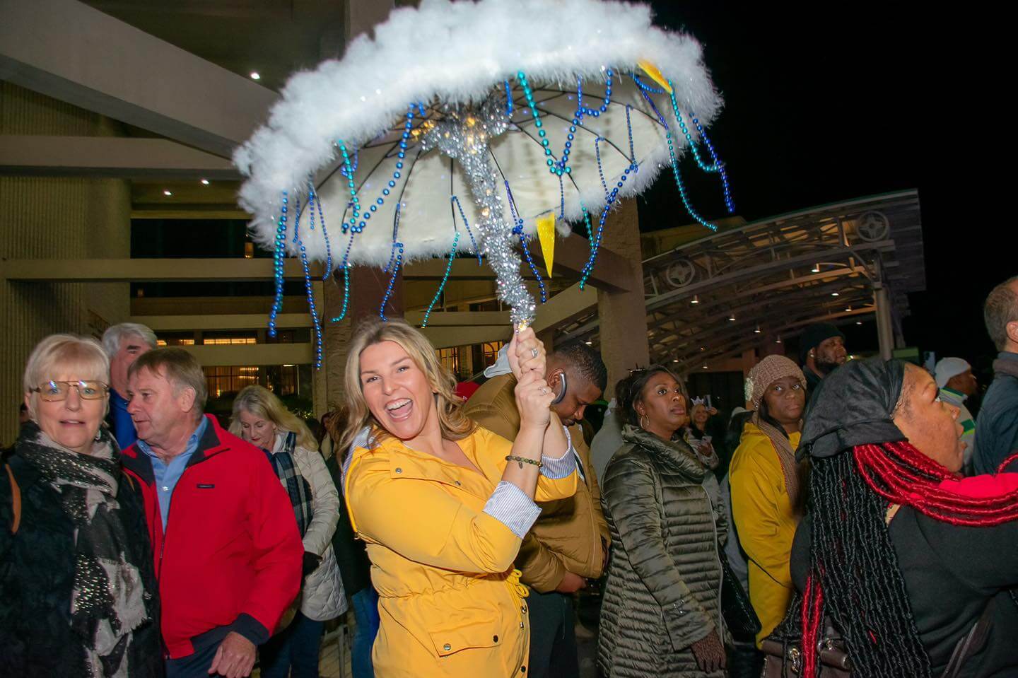 A woman in a yellow jacket holds an umbrella decorated with beads and fluff, standing amidst a crowd at the New Year's Eve countdown. Some wear jackets and hats, gathering together at this vibrant outdoor event as night envelops the city sky.