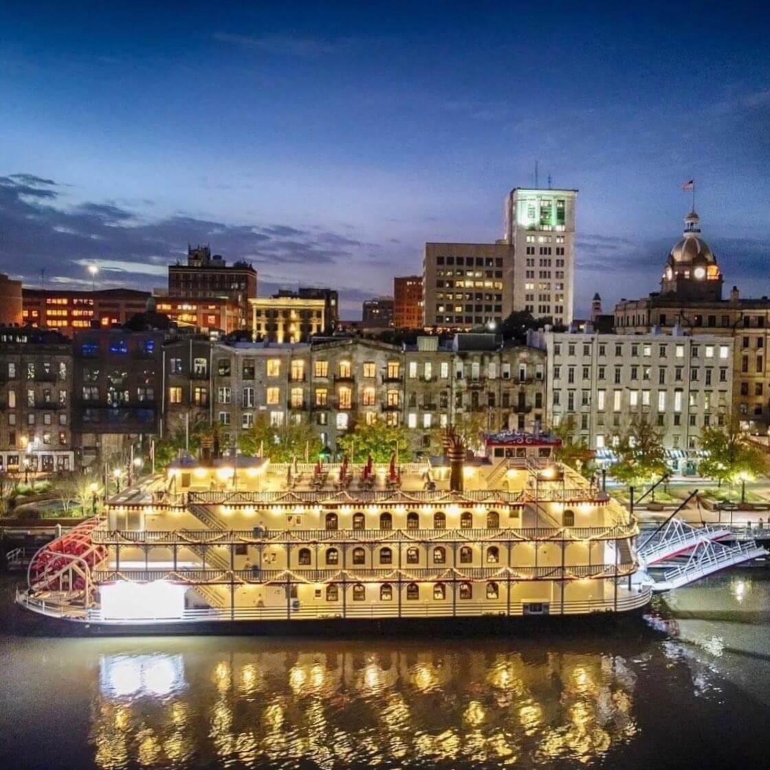 A lit-up riverboat docked at a city waterfront during the New Year's Eve countdown, with surrounding historic buildings glowing under the darkening sky.