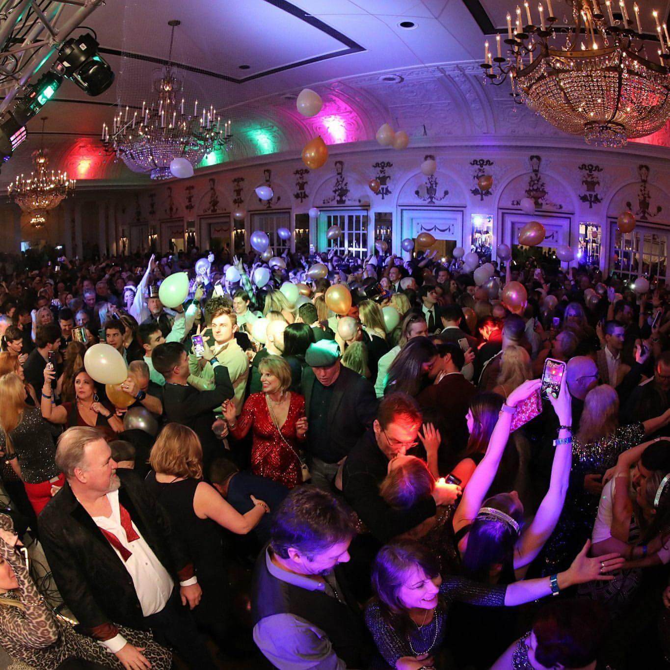 A large crowd gathered at a festive indoor event, celebrating the New Year's Eve countdown. People danced and held balloons under colorful lights and chandeliers, marking the joyous occasion with excitement in the air.