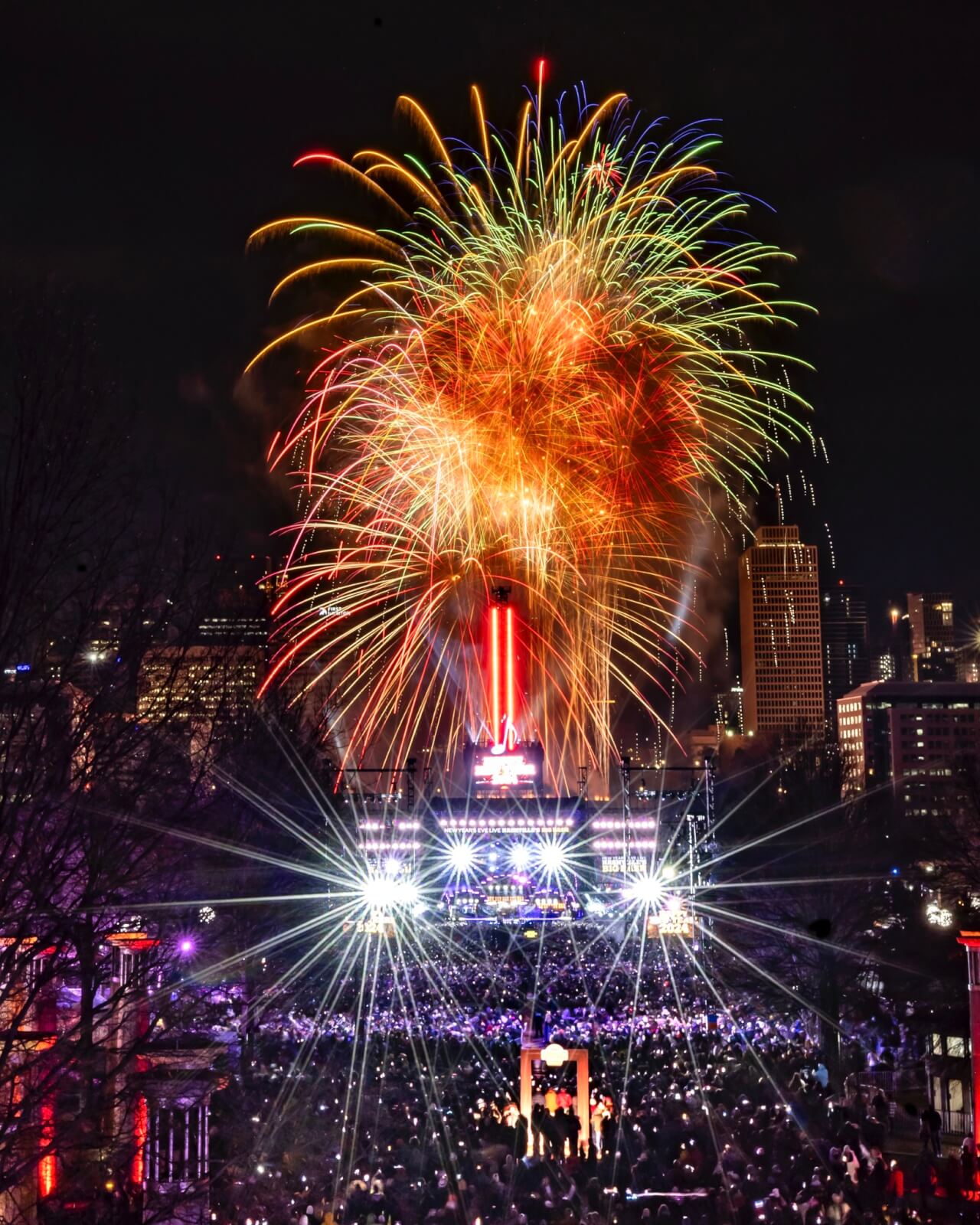 A large crowd eagerly gathers for the New Year's Eve countdown, watching a colorful fireworks display light up the city skyline at the nighttime outdoor event.