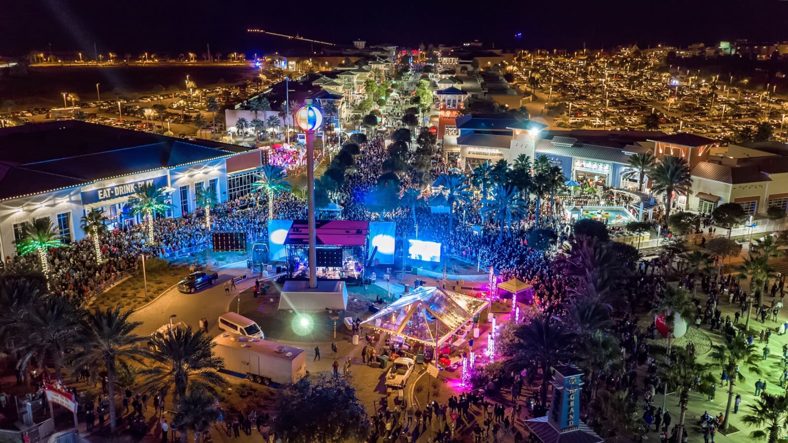 Aerial view of a bustling New Year's Eve festival in the South, with colorful lights, a large crowd, and various tents and stalls, surrounded by buildings and palm trees with a new year's eve countdown beach ball drop.