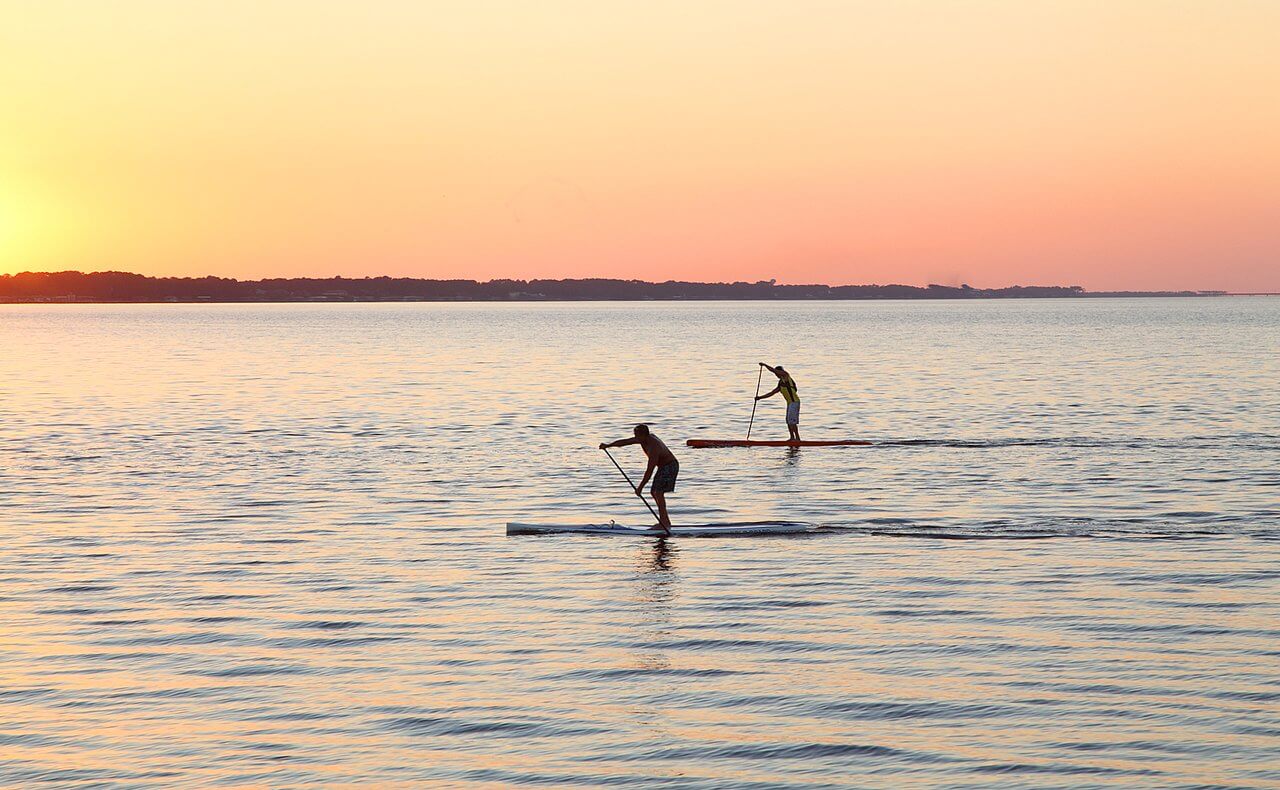 Two people paddleboarding on calm water during sunset, with an orange and pink sky, and distant land visible on the horizon.