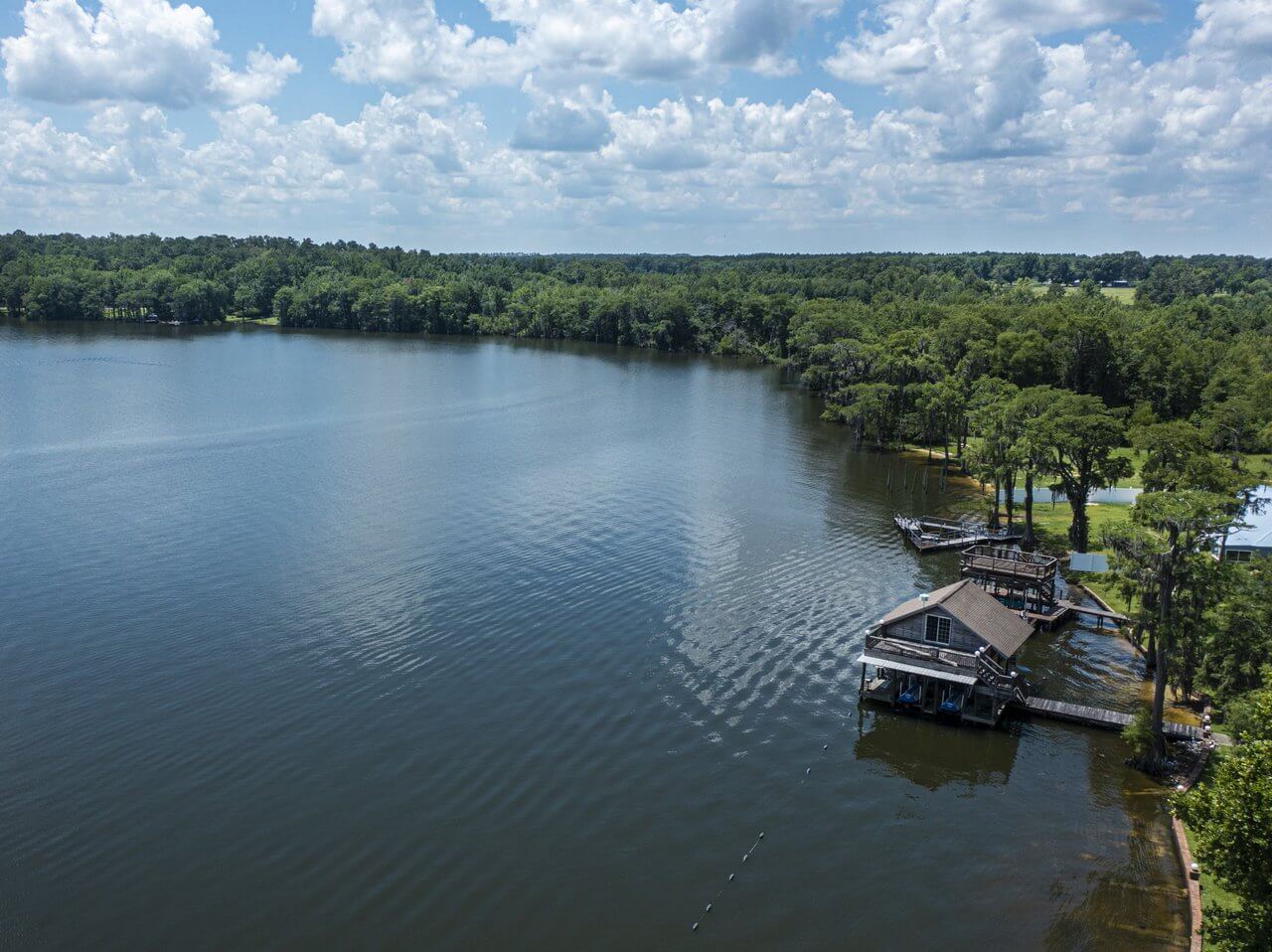 Aerial view of a serene lakeside with a dense forest in the background, a house on a dock to the right, and a cloudy sky overhead.