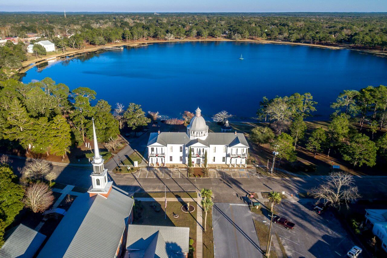 Aerial view of a large white building with a dome roof located near a calm lake, perfect for some Time on the Water, surrounded by trees. Adjacent buildings and roads are visible.