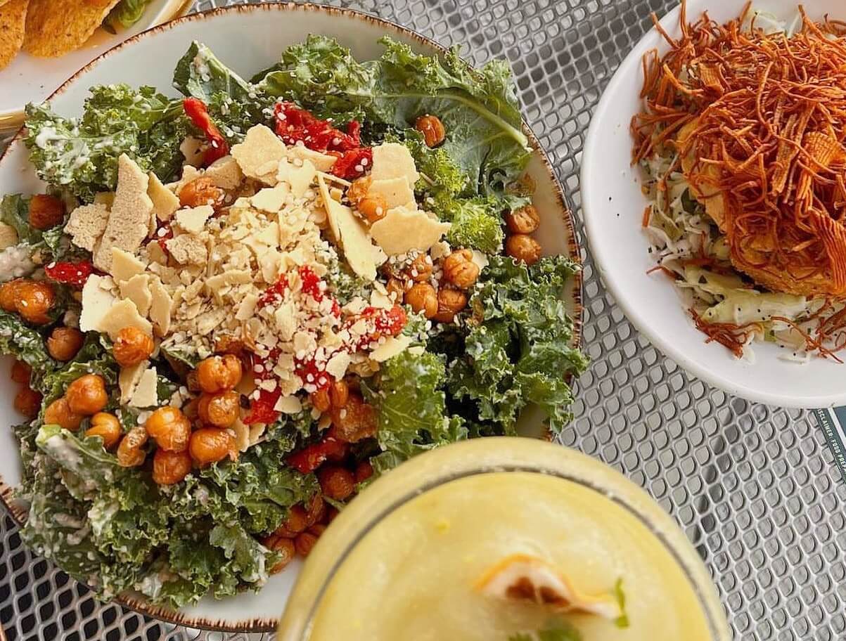 A bowl featuring one of the best salads in Nashville: kale salad with roasted chickpeas, shredded cheese, and red peppers sits alongside a dish of crispy noodles. In the foreground is a frothy yellow drink.