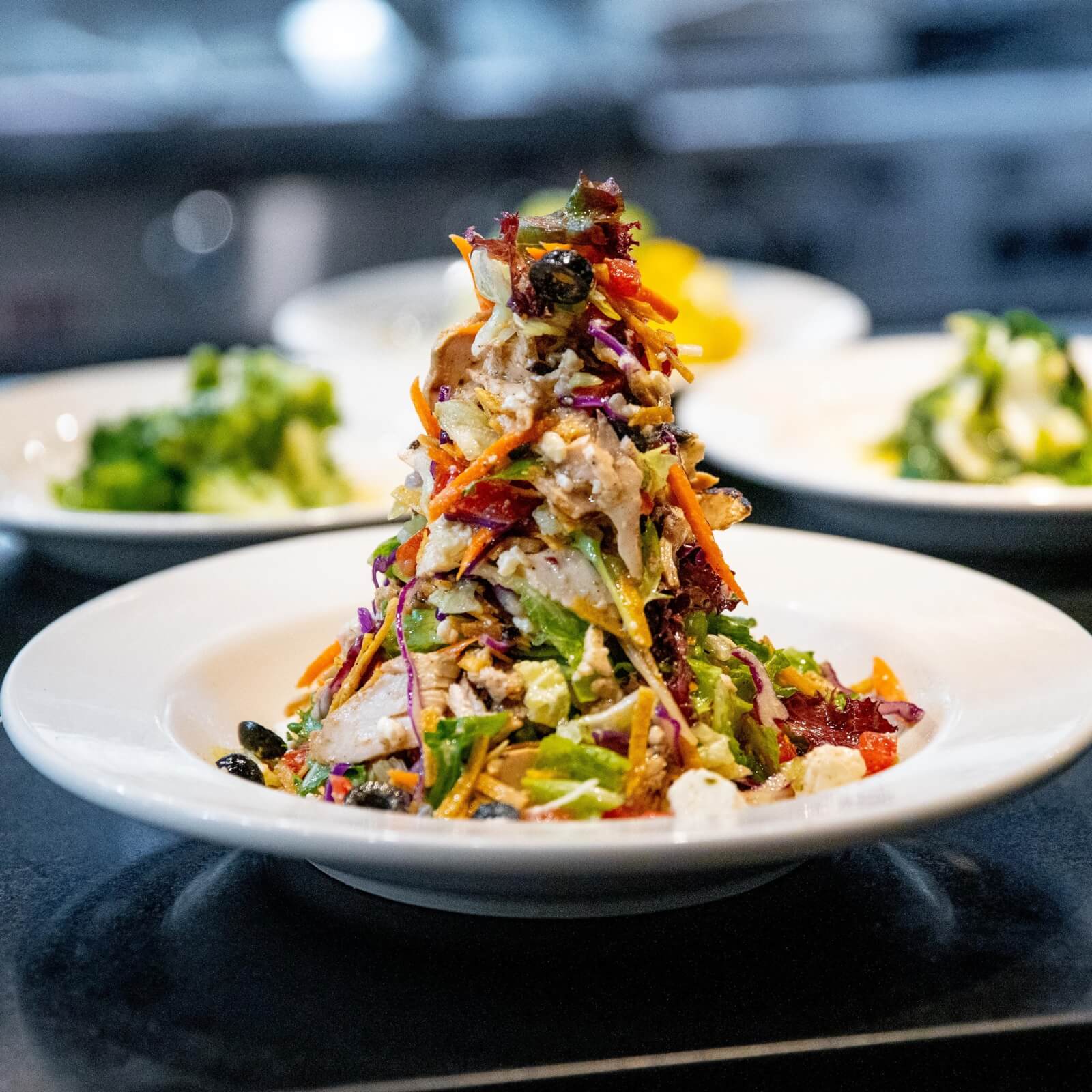 A colorful salad piled high on a white plate, featuring various vegetables and possibly chicken, captures the essence of some of the best salads in Nashville, with blurred plates of food in the background.