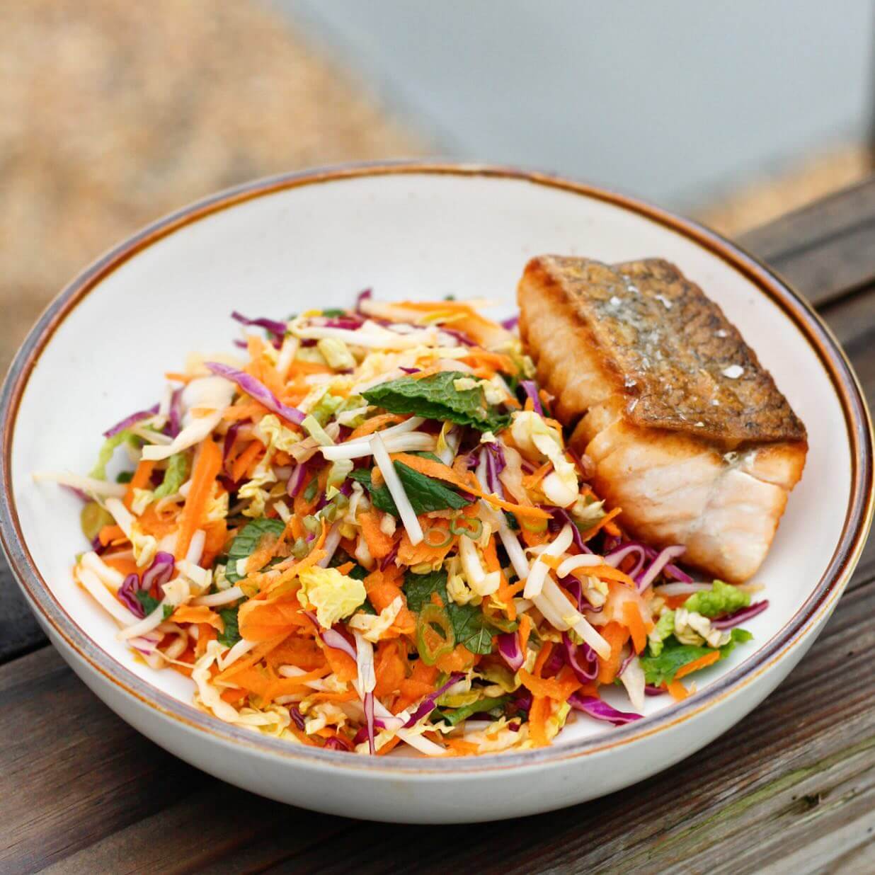 A bowl featuring salmon atop a vibrant vegetable salad with carrots, cabbage, and greens, all artfully arranged on a wooden surface.