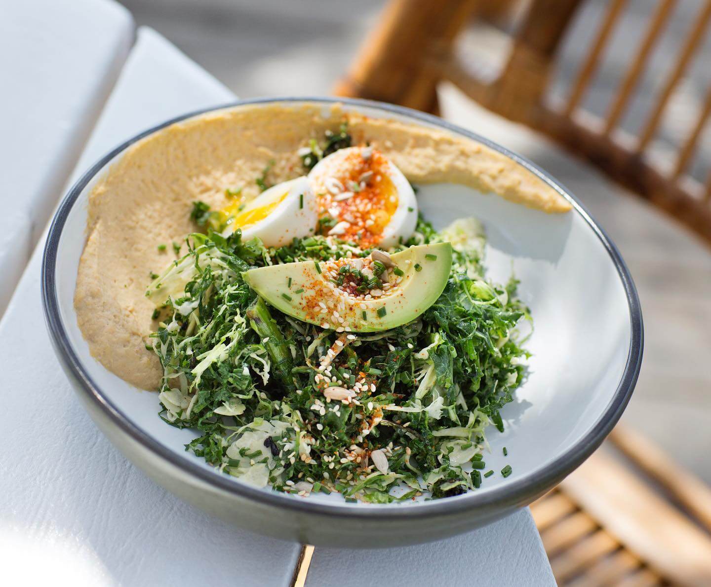 A bowl of salad with avocado slices, a halved boiled egg, chopped greens, and a spread of hummus, garnished with seasoning.