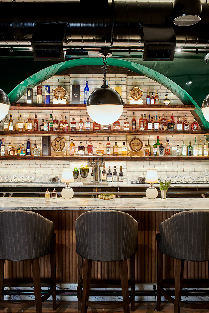 A modern bar interior in Birmingham restaurants showcases a marble countertop, striped cushioned bar stools, and a well-stocked glass shelf of assorted bottles beneath large spherical pendant lights.