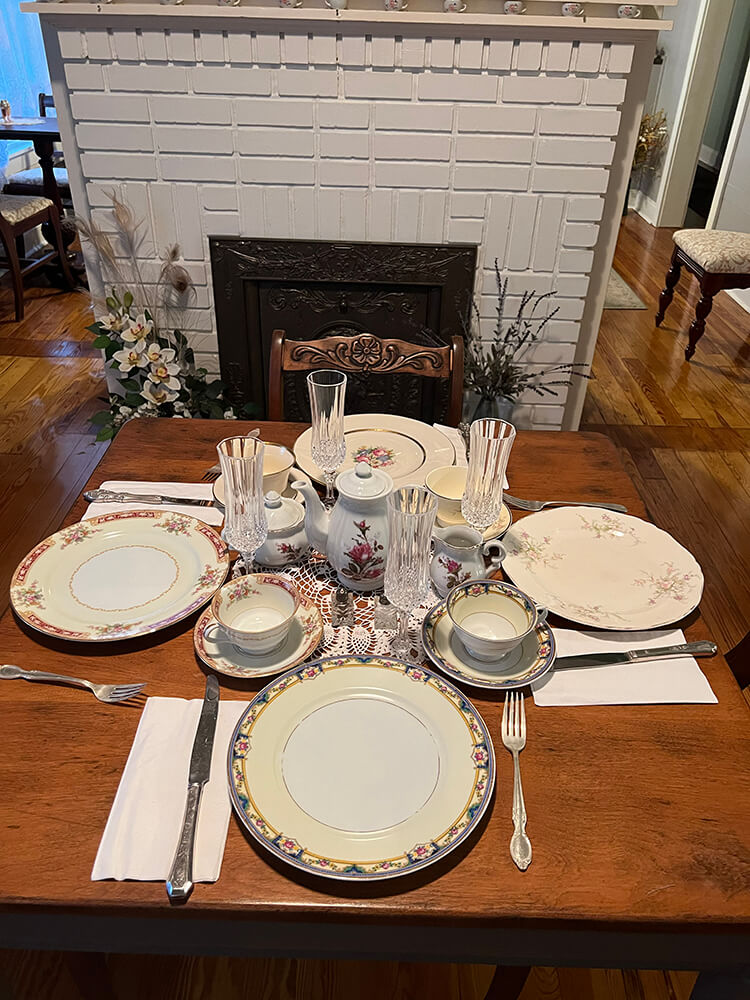 A dining table set for four with vintage plates, glasses, and a charming tea set ready for afternoon tea. The ambiance is completed by a white brick fireplace and decorative elements in the background.