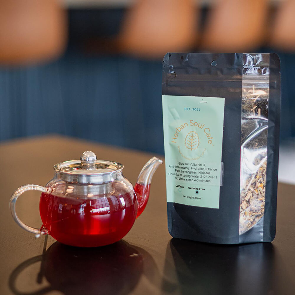 A glass teapot with red herbal tea sits elegantly next to a packaged herbal tea labeled "Haban Soul Cafe" on a dark table—an ideal addition to Birmingham's chic afternoon tea spots.