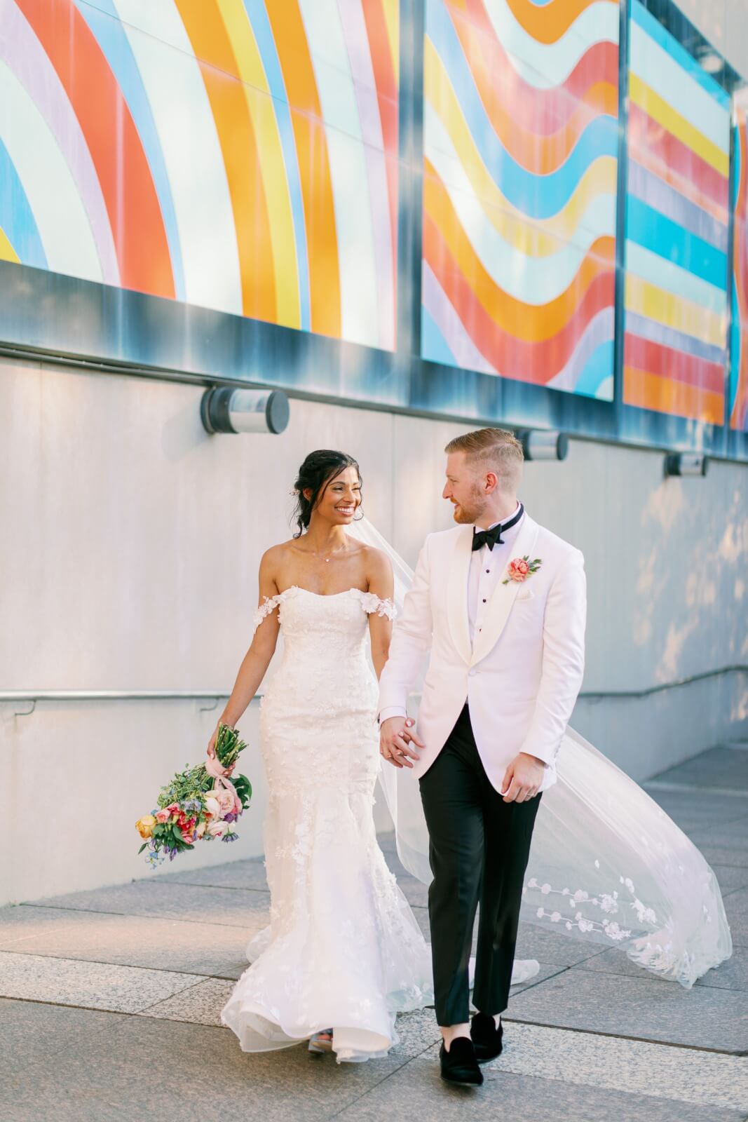 A bride and groom in formal wedding attire hold hands while walking outdoors past a colorful mural near the Birmingham Museum of Art. The bride carries a bouquet, and both are smiling.